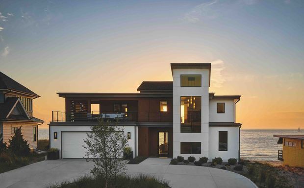 Exterior view of a contemporary multi-level coastal residence at sunset. The house combines crisp white rendered volumes with dark horizontal cladding, broad glazing, a projecting upper-level balcony, and a prominent narrow vertical window tower. A garage and driveway occupy the foreground, with neighbouring homes and the ocean visible beyond.