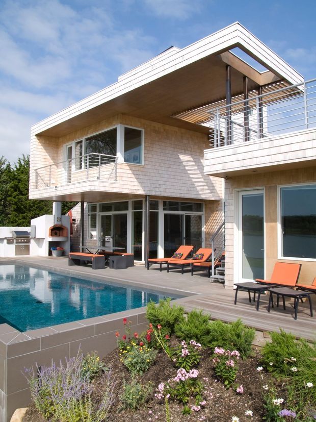 Exterior view of a modern multi-level residence surrounding a swimming pool. The home features light shingle-like cladding, extensive glazing, projecting balconies with horizontal metal railings, a deep roof overhang, timber soffits, and a pergola-like slatted terrace structure. Pool decking, outdoor lounge furniture, a built-in outdoor fireplace or oven, and landscaped planting occupy the foreground.