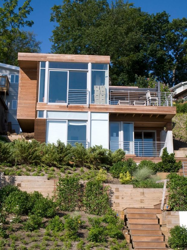 Exterior view of a contemporary multi-level residential home on a steep landscaped site. The facade combines warm horizontal timber cladding with extensive blue-tinted glazing, white-framed windows, and slender horizontal metal balcony railings. Multiple terraces and exterior stairs step down through timber retaining walls and planted garden beds.