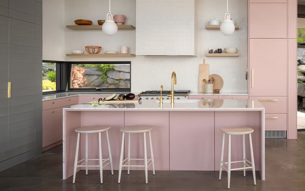 A refined contemporary kitchen with a large central island and four white stools. The cabinetry and island are dusty pink, complemented by a pale small-format tiled splashback, dark flooring, timber shelving, brass-toned tapware and hardware, and integrated appliances. Open shelving displays ceramics, while a wide window provides a view of a landscaped rock wall. The space is bright, polished, and softly colourful.