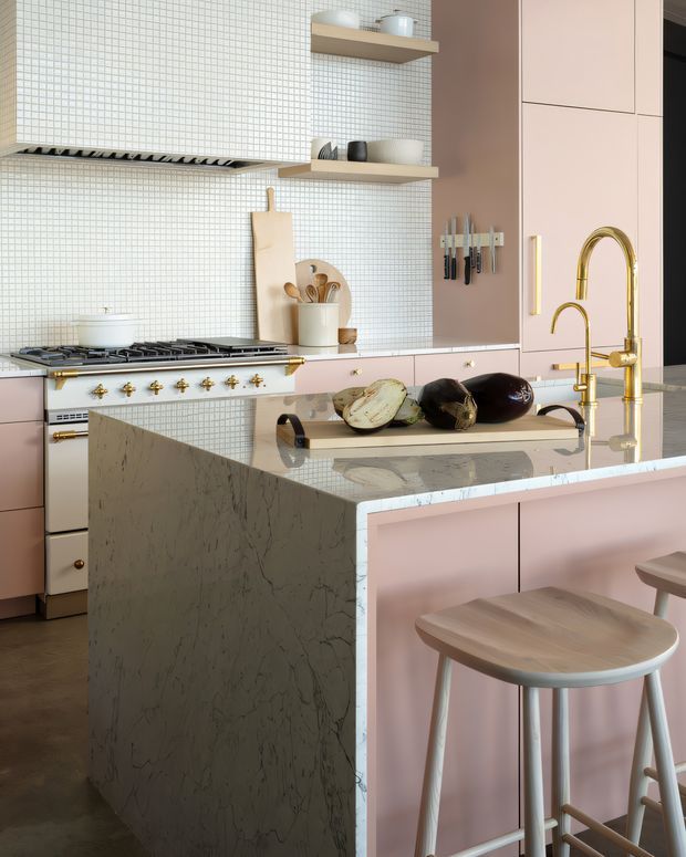 A refined pastel-toned kitchen featuring pink cabinetry, a large veined stone island with integrated seating, white micro-mosaic tiled walls, open timber shelves, brass fixtures and hardware, and a classic freestanding range. The composition includes a curved brass faucet, knife storage, cookware, and a wooden stool, creating a polished yet playful interior.