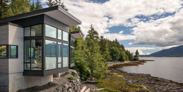 Exterior view of a contemporary, flat-roofed home projecting over a rocky waterfront shoreline. The building features dark horizontal cladding, light grey wall panels, extensive floor-to-ceiling glazing, and a concrete foundation, set among dense evergreen trees with mountains and open water beyond.