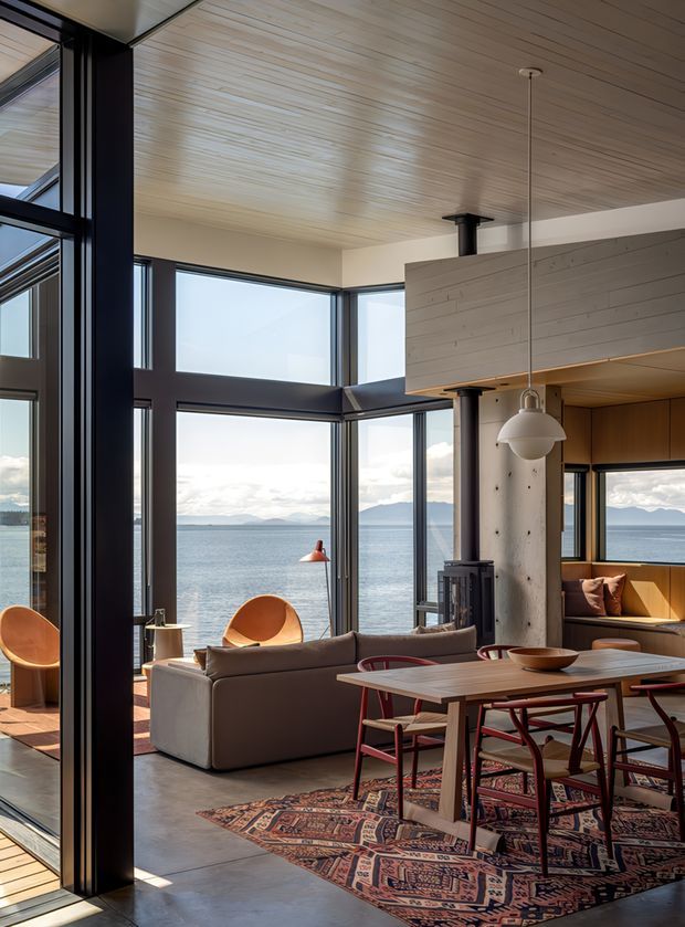 A warm, modern open-plan living and dining space with expansive floor-to-ceiling glazing overlooking a calm body of water and distant mountains. The room features a pale timber-lined ceiling, dark structural window framing, polished concrete flooring, a large patterned rug, a neutral sofa, sculptural lounge chairs, a timber dining table with red dining chairs, built-in timber seating, and a cylindrical concrete column with a wood-burning stove. A suspended globe pendant and abundant natural light complete the refined contemporary interior.
