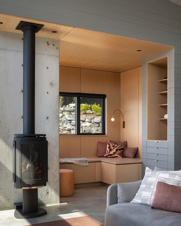 A compact, warmly detailed living room with a freestanding black wood-burning stove set against a raw concrete column. The room features extensive light timber wall and ceiling lining, integrated cabinetry, a built-in window seat, open shelving, and a grey sectional sofa with patterned and muted burgundy cushions. A black-framed window overlooks a landscaped stone retaining wall, while a slender brass wall light adds a small metallic accent. The design combines natural materials, built-in joinery, and contemporary cabin-like comfort.