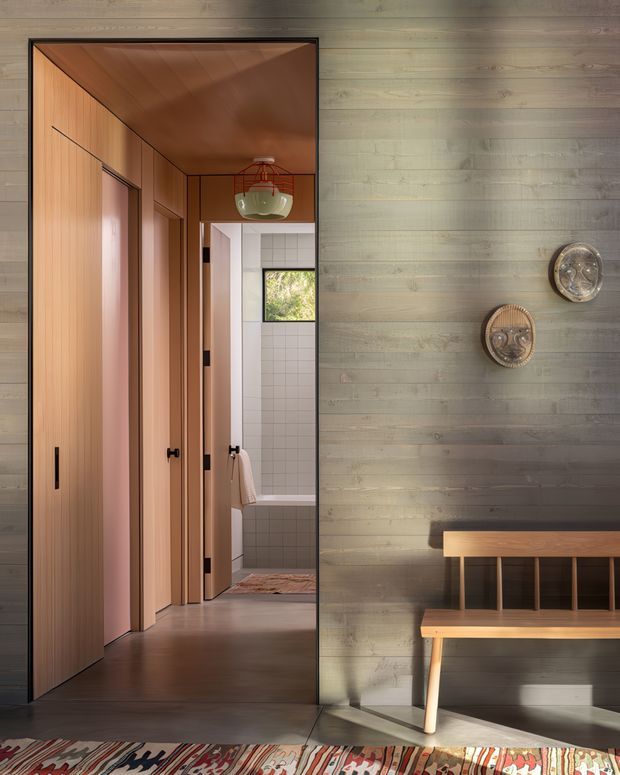 Interior view of a warm, timber-lined hallway and entry area. The composition features a large dark-framed opening, flush timber doors, a wood-paneled ceiling and walls, and a simple timber bench against a grey-green textured wall. A tiled bathroom is visible through the hallway, with decorative wall plates and a patterned rug adding detail.