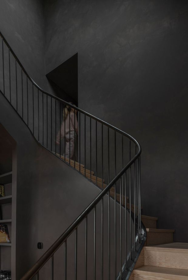 A dramatic interior view of a winding stairway with a curved black metal balustrade and slender vertical rods. The stair treads and risers are warm natural timber, contrasting with dark charcoal textured plaster walls. A person is partially visible on the upper flight, and a small section of built-in shelving appears at the lower left. The composition emphasizes the sculptural spiral-like geometry of the staircase and the moody, low-light atmosphere.