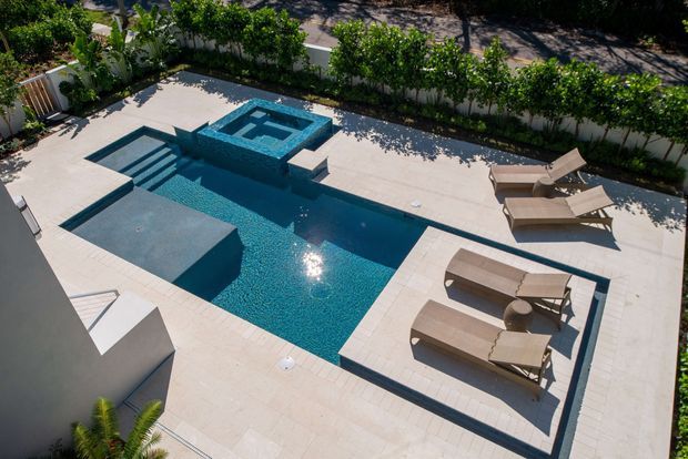 Aerial view of a modern landscaped pool courtyard featuring a geometric turquoise swimming pool with raised square spa or water feature, broad pale stone paving, built-in submerged platforms, and several tan sun loungers. Dense green planting and privacy screening border the space.