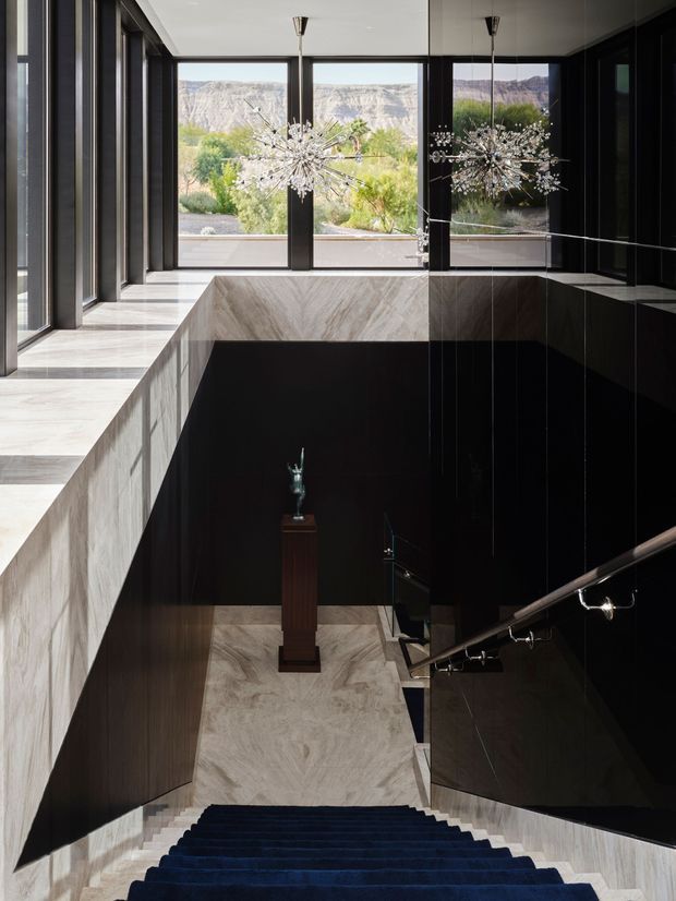 A dramatic modern stairway descends from a bright upper landing, framed by full-height dark window and mirror panels. Pale veined stone clads the landing edges and stair walls, while dark timber or black wall surfaces create strong contrast. Two sculptural starburst pendant lights hang above the stair void, with a small sculpture displayed on a central pedestal. Large windows overlook a dry, mountainous landscape.