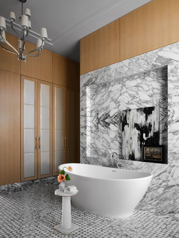 Elegant bathroom featuring a freestanding white bathtub, a wall-mounted chrome faucet, floor-to-ceiling white and grey veined marble wall cladding, grey mosaic tile flooring, and warm timber built-in cabinetry with mirrored inset panels. A classic multi-arm chandelier, artwork, and a small pedestal side table add decorative detail.