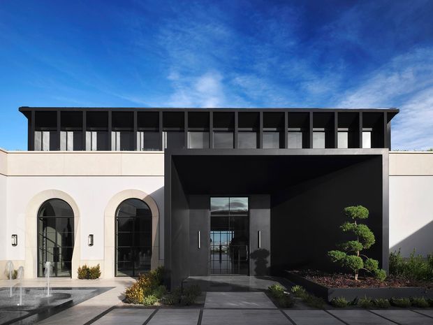Front exterior view of a sophisticated contemporary residence with a pale rendered facade, tall arched windows, and a dramatic black recessed entry portal. A dark upper level features a continuous rhythm of vertical framed glazing beneath a projecting flat roof edge. The entrance is approached by large square paved slabs, with a shallow reflecting pool and restrained landscaped planting including sculpted topiary. Bright blue sky and strong sunlight emphasize the crisp geometric composition.
