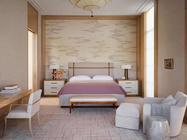 A refined bedroom with a centered upholstered bed, twin bedside tables and lamps, and a dramatic pale stone or marble-look feature wall framed by warm timber panelling. The room also includes a small dining or writing table with upholstered chairs, a pale lounge chair and ottoman, layered light carpeting, and a sculptural ceiling pendant. The palette is soft and warm, with blush, cream, beige, and light timber tones.