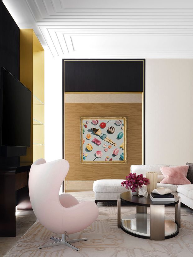 A refined living room with a sculptural pale pink lounge chair, cream sectional sofa, rounded black-and-metal coffee table, and textured area rug. The room features a dramatic black built-in media wall, warm timber feature panelling with brass trim, a large framed artwork, and an ornate white coffered ceiling. The palette is soft and neutral with black, gold, and pink accents.