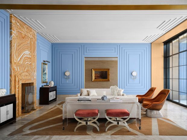 Luxurious, highly stylised living room with a symmetrical composition, blue geometric wall panelling, a veined warm-toned stone fireplace surround, white coffered ceiling, large black-framed glazing, and a marble-toned floor. A transparent dining table with red upholstered stools occupies the foreground, while a sofa, sculptural orange lounge chairs, framed artwork, and a television define the seating area. The room has strong Art Deco influences with polished, dramatic detailing.