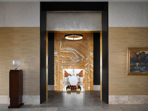 A refined interior hallway or entry passage framed by a dramatic black portal with brass detailing. The walls combine horizontally grained timber panelling with light stone wainscoting, while the floor is polished grey stone. A richly veined amber and cream stone feature wall leads to a secondary seating area, illuminated by a circular pendant. A dark timber pedestal with a glass ornament and a framed landscape artwork add decorative accents. The composition has a luxurious, architectural, Art Deco-inspired character.