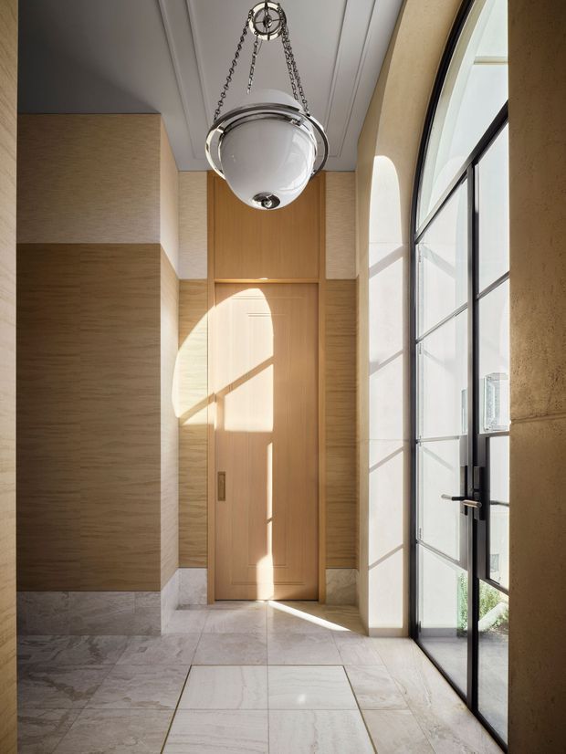 A refined, sunlit entryway with a tall paneled ceiling, pale natural timber wall panelling and a matching timber door. Large arched-top black-framed glazing and a glazed entry door line the right side, while pale veined stone tiles cover the floor. A chrome-and-glass pendant light hangs centrally, and strong sunlight creates geometric shadows across the door and floor.