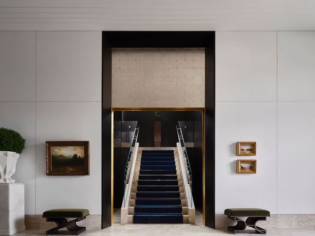 Formal interior view of a grand stairway framed by a black and brass-toned architectural portal. The staircase has dark carpeted treads, pale stone or marble side walls, glass balustrades, and illuminated handrails. Large pale wall panels, a light coffered or paneled ceiling, framed artwork, sculptural decor, and low dark benches flank the stair. The composition is symmetrical, refined, and hotel-like.