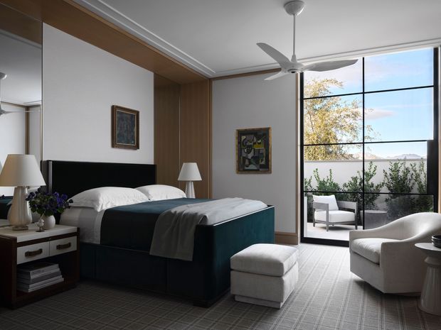 A refined contemporary bedroom with a dark upholstered bed, white bedding, bedside tables and lamps, a matching upholstered ottoman, and a curved lounge chair. Warm timber wall panelling and built-in cabinetry frame the bed, while full-height black-framed glazing opens to an outdoor seating area. A ceiling fan, artwork, layered neutral textiles, and patterned carpet complete the room.