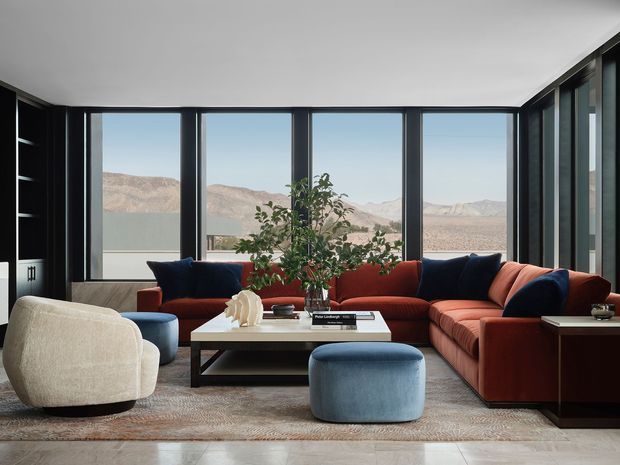 A refined contemporary living room with a large sectional sofa arranged around a low rectangular coffee table. Floor-to-ceiling black-framed glazing spans the rear and side walls, offering expansive views of a dry mountainous landscape. The room includes sculptural upholstered lounge chairs, a blue ottoman, dark built-in shelving, a neutral area rug, and a small indoor tree. The palette combines warm rust-red upholstery with deep navy, cream, charcoal, and natural timber tones.