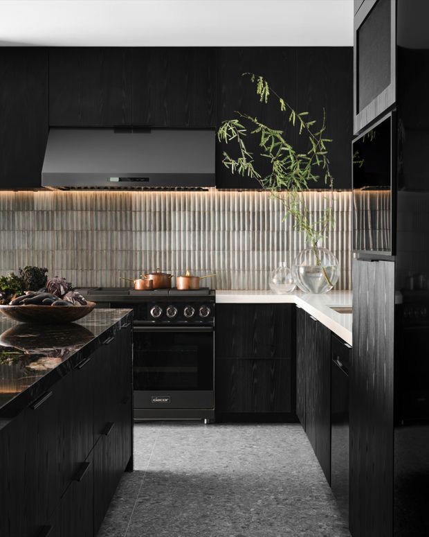 A moody, compact contemporary kitchen with black vertically grained cabinetry, a dark stone island benchtop, a pale stone perimeter benchtop, a sculptural ribbed grey backsplash, integrated rangehood, freestanding black cooker, and warm under-cabinet lighting. A narrow internal layout is softened by glass vessels and greenery.