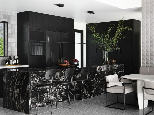 A sophisticated contemporary kitchen and dining area featuring dramatic black cabinetry, a large black-and-white veined stone island and matching stonework, black bar stools, integrated appliances, and a white dining table with upholstered chairs. Textured geometric wall tiles and sculptural pendant lights add visual interest, while greenery and monochrome finishes create a refined, high-contrast interior.
