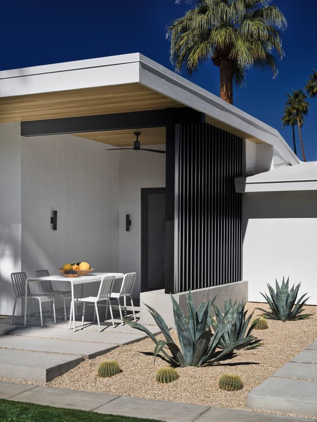 Exterior view of a low-slung modern residence with a flat roof, crisp white rendered walls, dark vertical screening, and a warm timber-lined covered patio. A small outdoor dining setting sits beneath the overhang, with gravel landscaping, agave and barrel cacti, palms, and concrete paving creating a desert-modern character.