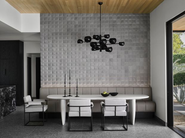 A sophisticated dining area with a long oval dining table, upholstered banquette seating, modern armchairs, and a sculptural black multi-arm pendant light. The room features a dramatic three-dimensional geometric tiled feature wall, a warm timber ceiling, dark cabinetry or wall finishes, and large glazed doors opening to an outdoor garden area.