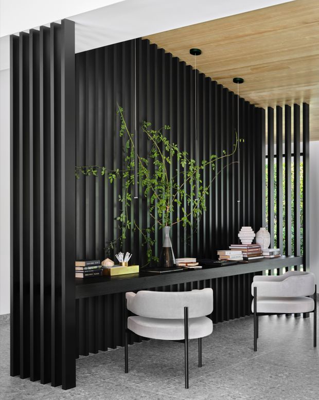 A refined contemporary interior featuring a dramatic black vertical slatted partition and integrated floating console shelf. Two light upholstered lounge chairs sit on a grey stone-look floor, while a warm timber-lined ceiling and greenery-filled decorative display add contrast and softness. Narrow openings in the slatted wall provide filtered views toward greenery outside.