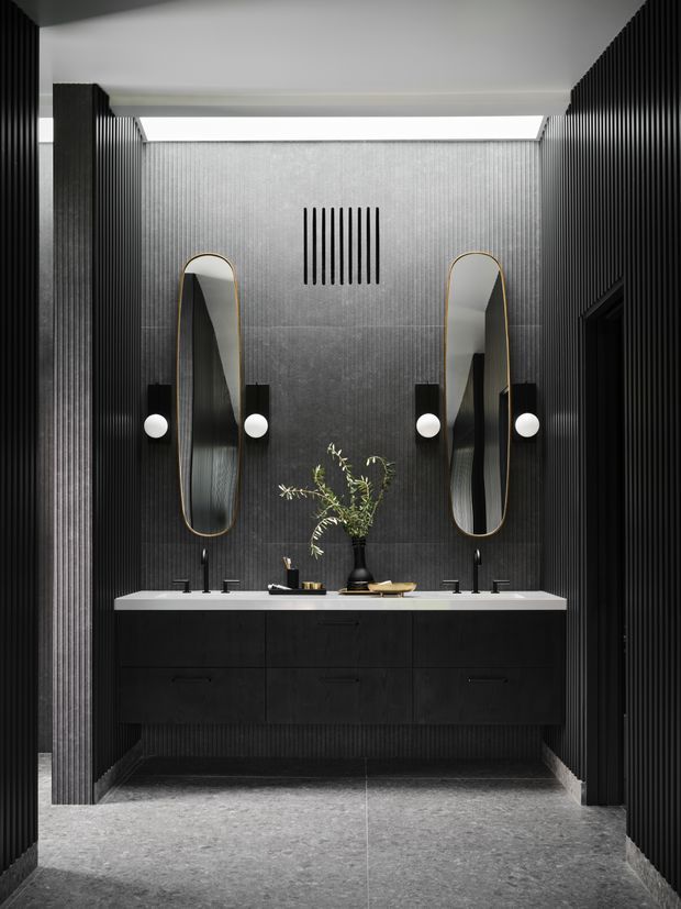 A dramatic, symmetrical contemporary bathroom vanity with a double basin, floating dark cabinetry, pale stone benchtop, tall arched mirrors with brass-toned frames, globe wall lights, and black fluted wall panelling. The space has a textured grey feature wall, a linear skylight or clerestory above, and a grey stone-look floor.
