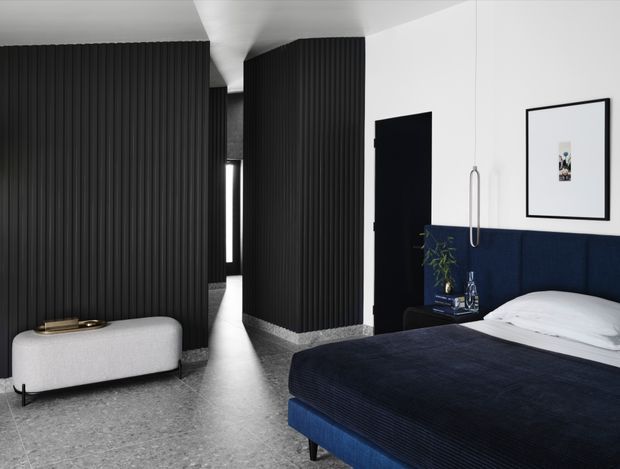 Contemporary bedroom with a dark navy upholstered bed, white bedding, black vertically ribbed wall panelling, and a white wall with framed artwork. A sculptural light-grey bench sits on a polished grey floor, while a narrow passage and black doorway create a strong graphic contrast.