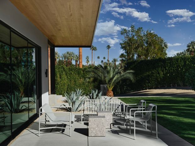 Modern outdoor terrace beside a glazed living area, with a broad timber-lined overhang, concrete paving, white metal lounge and dining furniture, and a landscaped garden screened by tall hedges and palms. The setting is bright and relaxed, with strong indoor-outdoor connection.