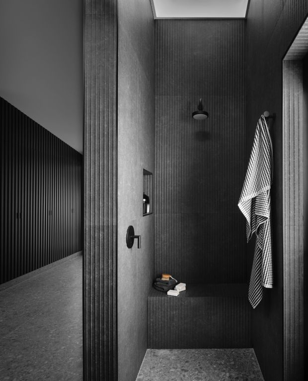 Moody monochrome architectural photograph of a compact walk-in shower with dark vertically ribbed wall cladding, a black shower fixture, recessed niche, skylight, towel hooks and a stone-look floor. An adjoining dark ribbed corridor or passage is visible to the left.