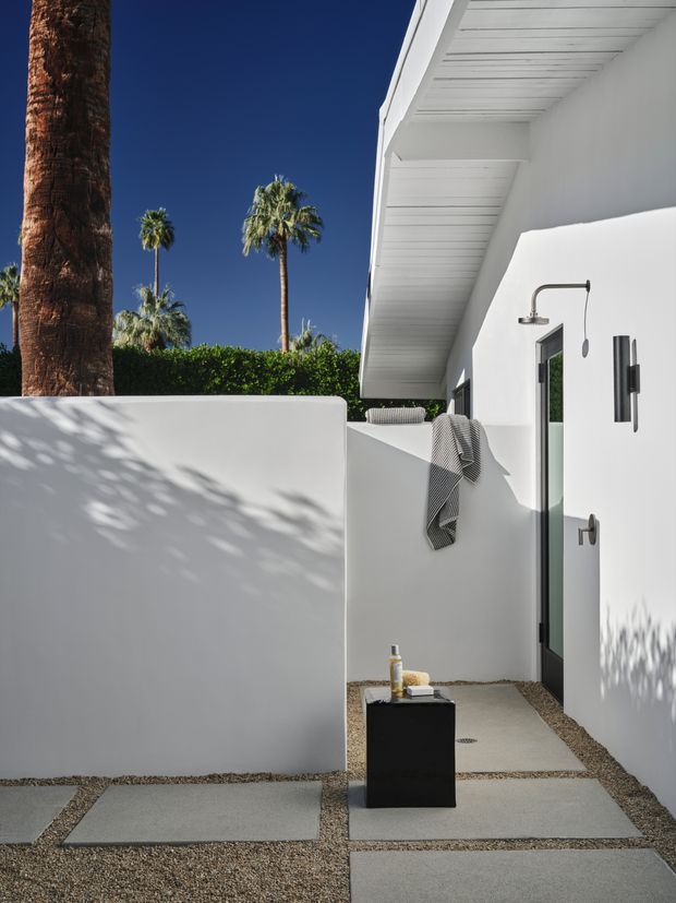 Minimalist white outdoor shower courtyard beside a modern flat-roofed building, with large concrete stepping slabs set into a gravel surface, a black pedestal holding toiletries, and a wall-mounted shower fixture. Palm trees and deep blue sky are visible beyond the privacy walls.