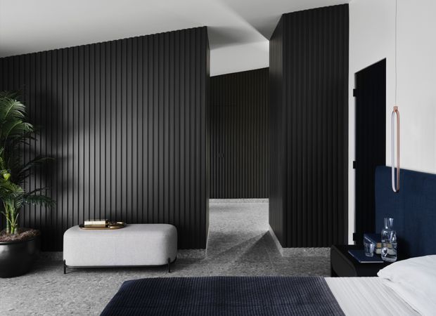 A sophisticated modern bedroom featuring dramatic black vertical fluted wall panelling, a low light-grey upholstered bench, dark textured flooring with a navy rug, and a navy upholstered headboard. A minimalist black bedside table with glassware sits beside the bed, while a tall indoor plant adds greenery. The room has a restrained monochrome palette with deep blue accents and strong linear detailing.