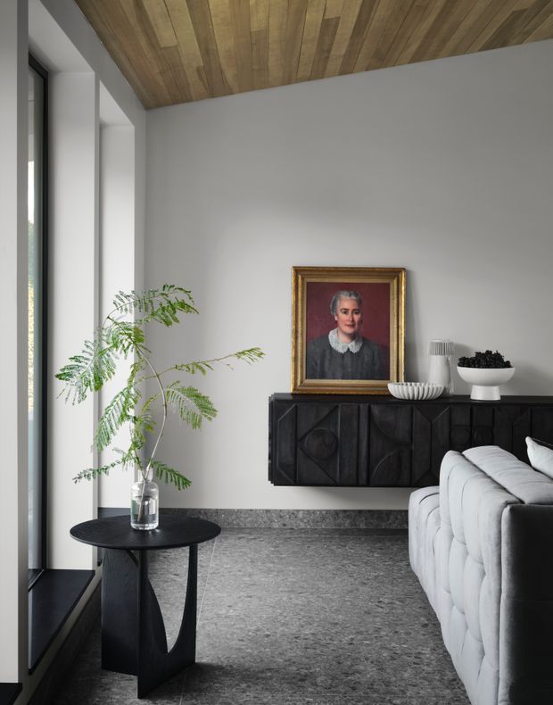 A refined contemporary living room with a large light-grey upholstered sofa, a black sculptural side table, and a wall-mounted black geometric timber sideboard. The room features a polished grey stone floor, white plaster walls, a warm timber-lined ceiling, tall dark-framed glazing, and a framed portrait with decorative objects on the sideboard. A leafy branch in a glass vase adds an organic accent.