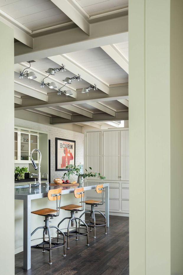 A bright, refined kitchen viewed through a wide opening, featuring a long grey island with integrated sink and three industrial-style timber-and-metal stools. The room has pale green-grey cabinetry, white brick walls, dark timber flooring, and a highly articulated white-painted coffered and exposed-beam ceiling with multiple adjustable pendant-style fixtures. A poster, plants, open shelving, and kitchenware add character.