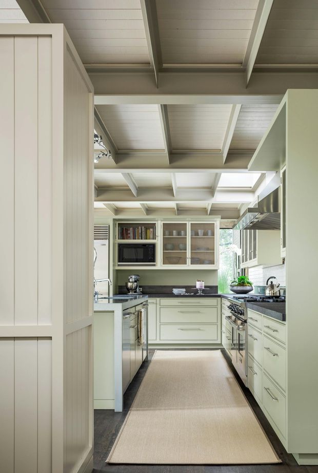 A bright, compact galley-style kitchen viewed through a partially enclosed entry. The kitchen features pale sage-green shaker cabinetry, dark stone-looking benchtops, stainless-steel appliances, glass-front upper cabinets, and a long neutral runner rug. The room is defined by a white-painted, tongue-and-groove ceiling with exposed boxed beams and a skylight, creating a light, tailored traditional interior.