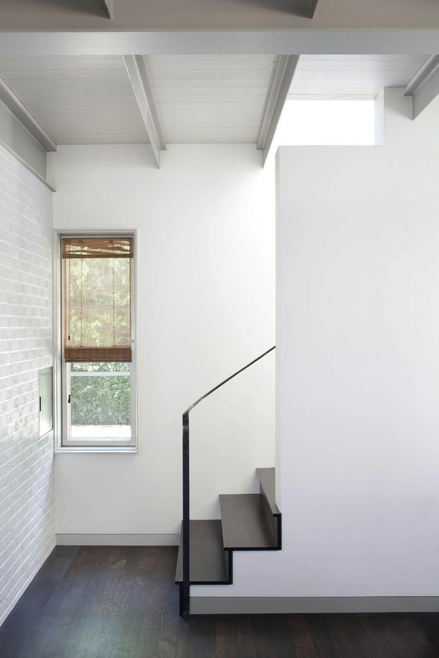 Bright, minimalist stairway interior with white-painted brick and plaster walls, exposed white ceiling beams, dark timber flooring, and a slim black metal stair balustrade. A narrow window with a woven timber blind brings in filtered natural light. The compact stair turns around a white central wall, creating a clean, sculptural composition.