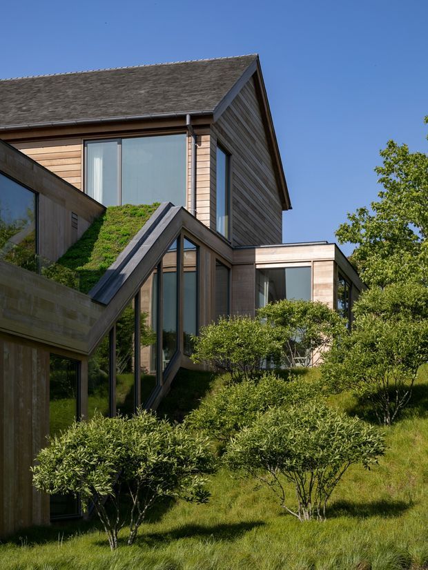 A contemporary multi-level residential building set within a landscaped, grassy garden. The structure features a steep gabled roof, extensive floor-to-ceiling glazing, horizontal timber cladding in natural brown tones, and angular projecting volumes, including a green-roofed section. Mature shrubs and trees partially screen the lower levels.
