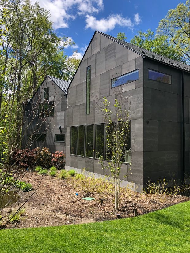 Exterior view of a contemporary dark-clad building set within a wooded landscaped garden. The structure has steep gable roofs, large rectangular windows, and a minimalist facade formed from dark grey panel cladding, with a smaller connected gabled volume visible behind it.