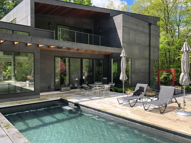 A contemporary multi-level house opening onto a paved pool terrace. The building has dark charcoal panelled walls, extensive glazing, a projecting flat roof, a cantilevered upper balcony with slim metal railings, and warm timber soffits. The foreground contains a rectangular swimming pool, outdoor dining furniture, loungers, and closed umbrellas, surrounded by mature greenery.