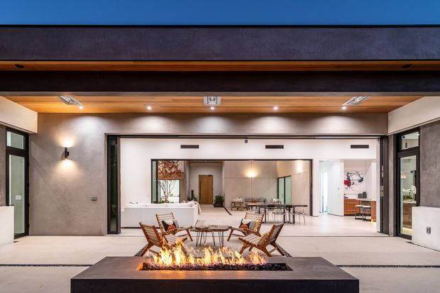 A wide, open-plan contemporary indoor-outdoor entertaining area viewed from a covered terrace. The space features a long linear fireplace in a dark stone or concrete plinth, a dining table with woven chairs, warm timber-lined ceiling, expansive dark-framed glazed openings, and a bright living and dining area beyond. Neutral plastered walls and integrated lighting create a refined, warm atmosphere.