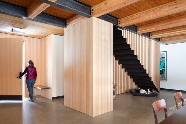 Open-plan contemporary interior featuring a timber-lined ceiling with exposed dark steel beams, extensive vertical timber wall panelling, a sculptural dark staircase with timber balustrade, polished concrete flooring, and part of a dining area in the foreground.