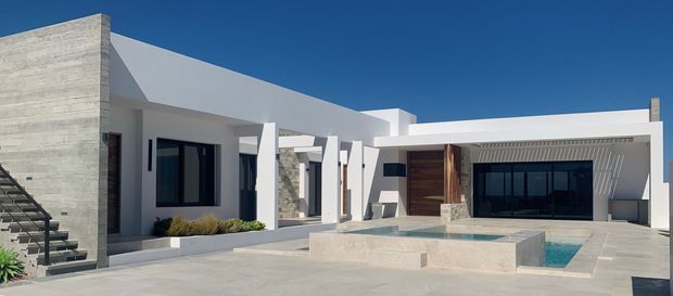 A low-slung contemporary residence arranged around a paved courtyard and rectangular swimming pool. The building has crisp white rendered walls, strong geometric volumes, a flat roofline, extensive dark-framed glazing, a warm timber-clad entry volume, and a prominent textured concrete feature wall. Exterior stairs and minimalist landscaping complete the bright, resort-like composition under a clear blue sky.