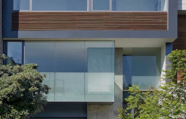 A close exterior view of a contemporary multi-level building facade, featuring extensive blue-grey glazing, a projecting glass balcony or enclosure, dark vertical wall panels, smooth concrete surfaces, and a horizontal timber-look louvred screen beneath the upper windows. Landscaping partially frames the lower edges.