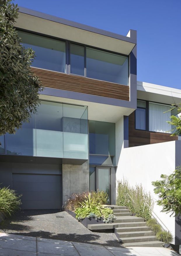 Exterior view of a contemporary multi-storey urban residence with a flat roof, strong geometric volumes, dark metal-framed glazing, frosted glass balcony balustrades, horizontal timber screening, rendered walls, and a concrete-clad entry zone. A garage is visible at ground level, with a landscaped stairway leading to the entrance.