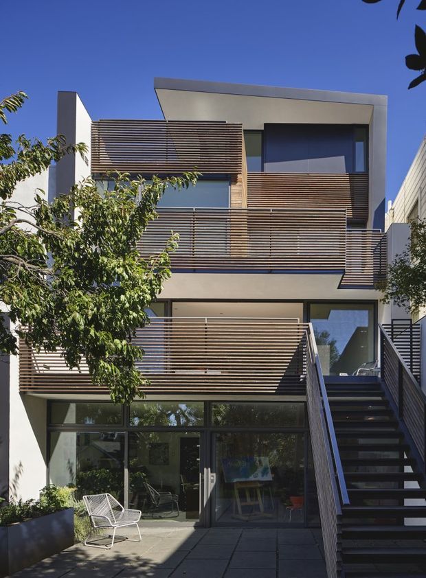 Front exterior view of a narrow multi-storey contemporary residence with layered balconies, horizontal timber privacy screens, white rendered walls, dark-framed glazing, and an external stair rising beside a paved courtyard or entry terrace. The facade has strong horizontal bands and recessed openings, with mature planting partially screening the lower level.