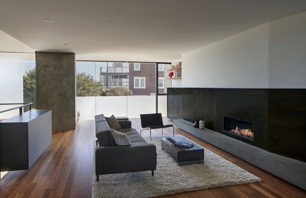 A bright, open-plan contemporary living room with full-height glazing, timber flooring, a charcoal sectional sofa, lounge chair, coffee table, and textured rug. A dramatic dark stone or concrete fireplace wall with a built-in fire forms the main feature, while a minimalist kitchen island is visible at the left.