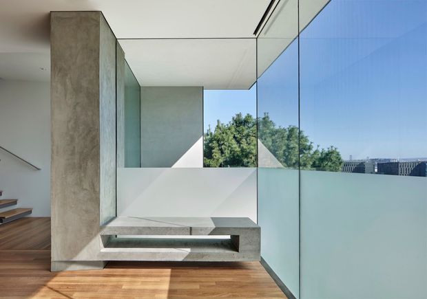 A bright contemporary interior landing or living area with floor-to-ceiling glazing overlooking trees and rooftops. A monolithic concrete feature structure incorporates a low built-in bench or platform beside a tall concrete wall, while warm timber flooring and crisp white ceilings and walls create a clean, minimal composition. A stairway is partially visible at the left edge.