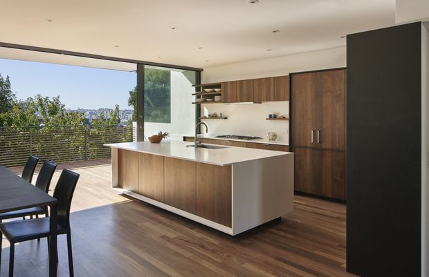 Contemporary open-plan kitchen and dining area with a large white island, integrated sink and tap, dark timber cabinetry, white benchtops, and warm timber flooring. Floor-to-ceiling sliding glazing opens to a timber deck and leafy urban outlook. A dining table and chairs occupy the foreground.