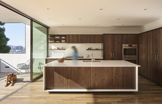A bright, spacious contemporary kitchen with a large central island, dark timber cabinetry, integrated appliances, open shelving, and a pale stone benchtop. Full-height glazed sliding doors connect the kitchen to an exterior terrace or balcony, with a city view beyond. Warm timber flooring and abundant daylight create an inviting, refined atmosphere.
